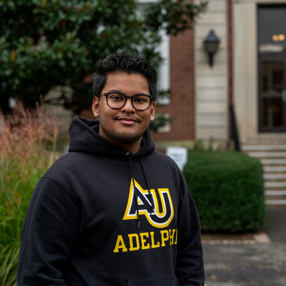 A confident male Ҵý student stands in front of Levermore Hall wearing an Adelphi hoodie.