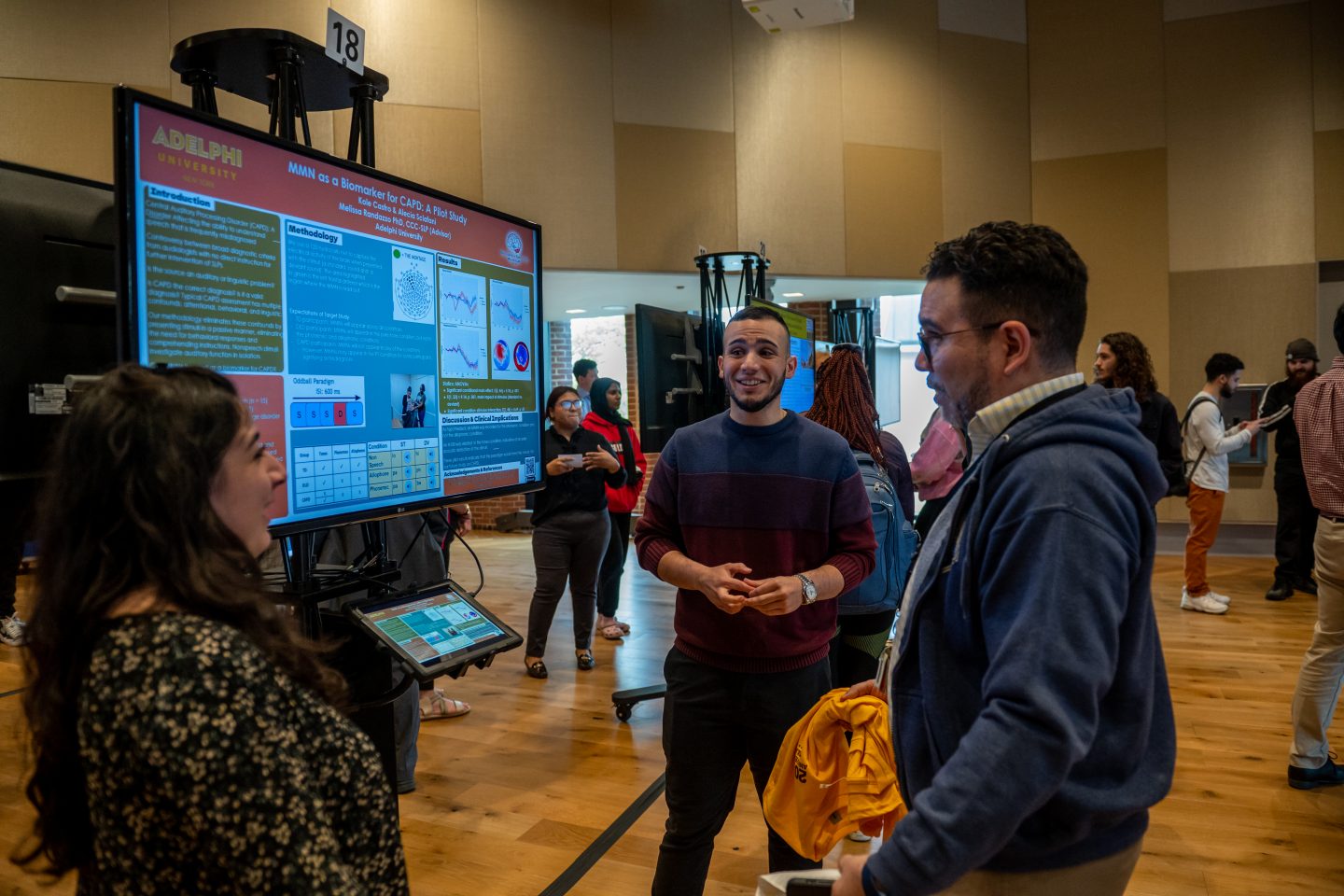 Three Adelphi students standing by an e-poster discuss a 2023 Research Day presentation on biomarkers in the University Center.